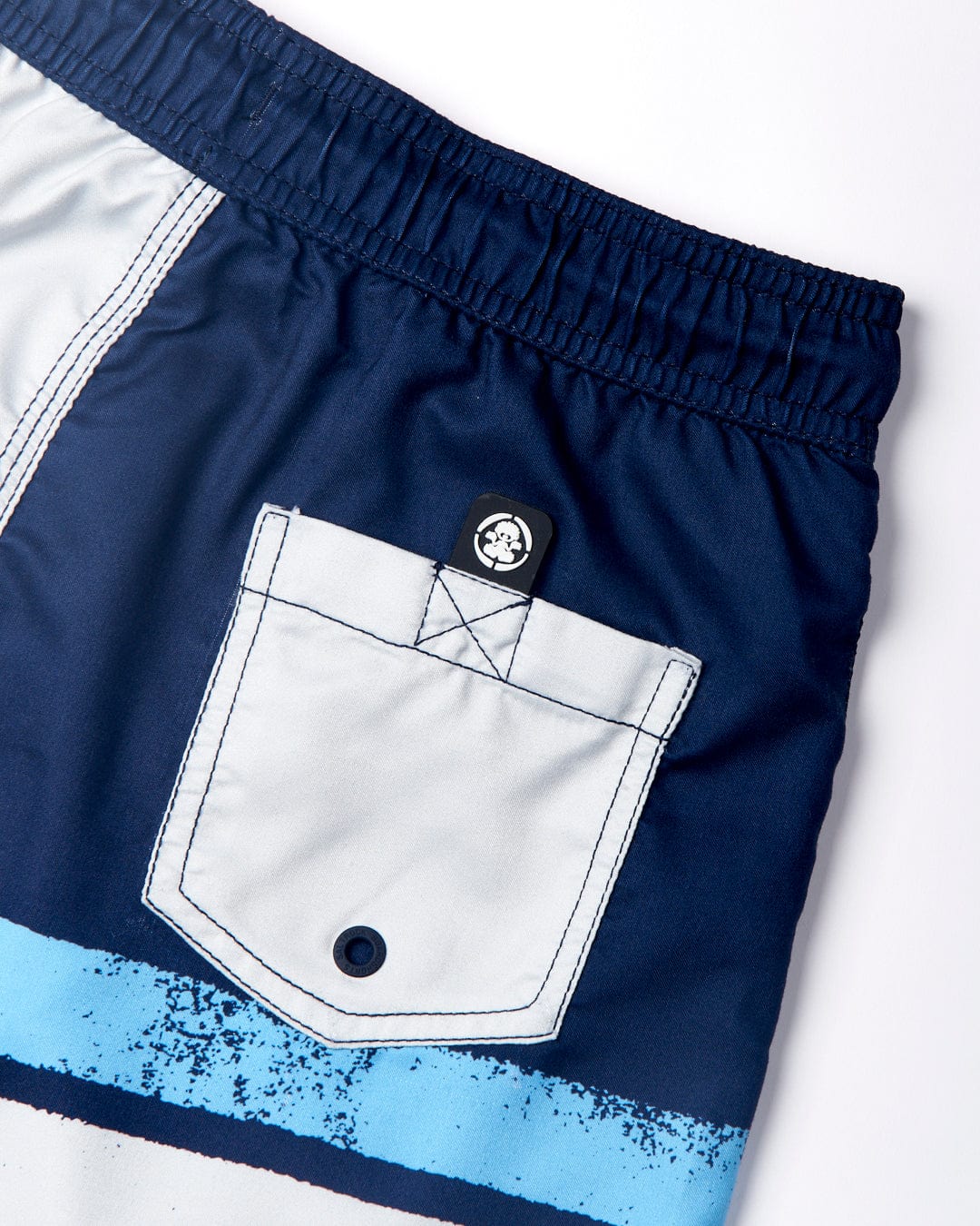 Close-up of a pair of Saltrock Original SR - Kids Swimshorts - Blue with an elasticated waist and a white back pocket featuring a button. Sporting two light blue horizontal stripes at the lower part, these swimshorts also showcase branded dipped rubber end drawstrings for added style.