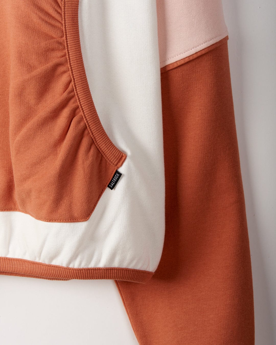 Close-up of a color-blocked garment featuring white, light pink, and rust orange cotton fabric. There is a small black tag with Saltrock branding attached near the seam, highlighting its loose fit. This piece is the Palmera - Womens Pop Hoodie - Cream by Saltrock.