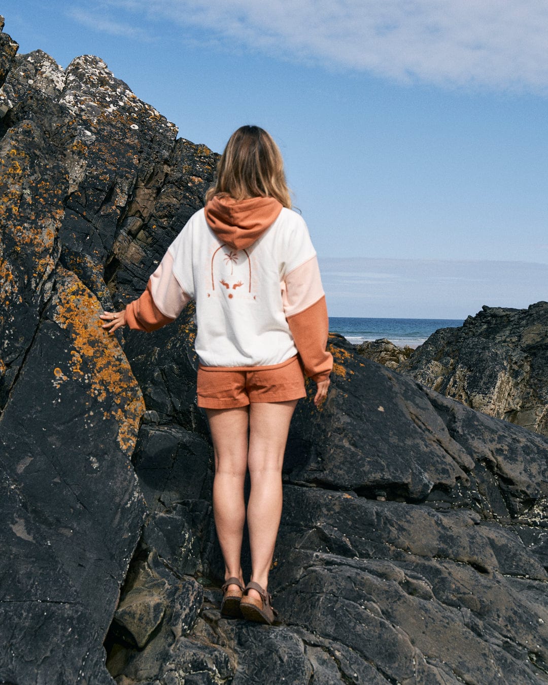 A person in a Palmera - Womens Pop Hoodie - Cream with Saltrock branding stands on rocky terrain, facing away towards the ocean, their loose fit shorts fluttering in the breeze.