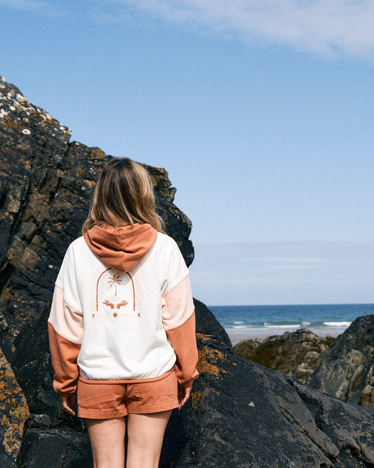 Person wearing a **Palmera - Womens Pop Hoodie - Cream** and shorts, standing on rocky terrain with the ocean in the background. The cotton material hoodie has a palm tree design on the back and **Saltrock** branding.