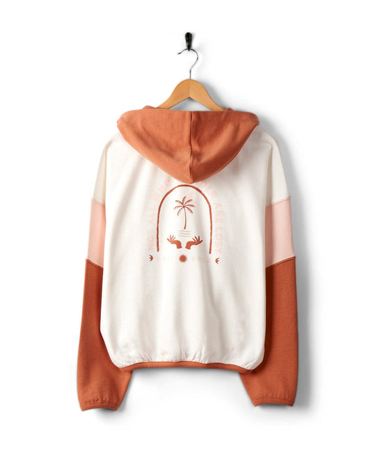 A Palmera - Womens Pop Hoodie - Cream by Saltrock with a white and orange color block design, hanging on a wooden hanger. Made from 100% cotton, the back features a circular palm tree graphic with text and two hand illustrations.