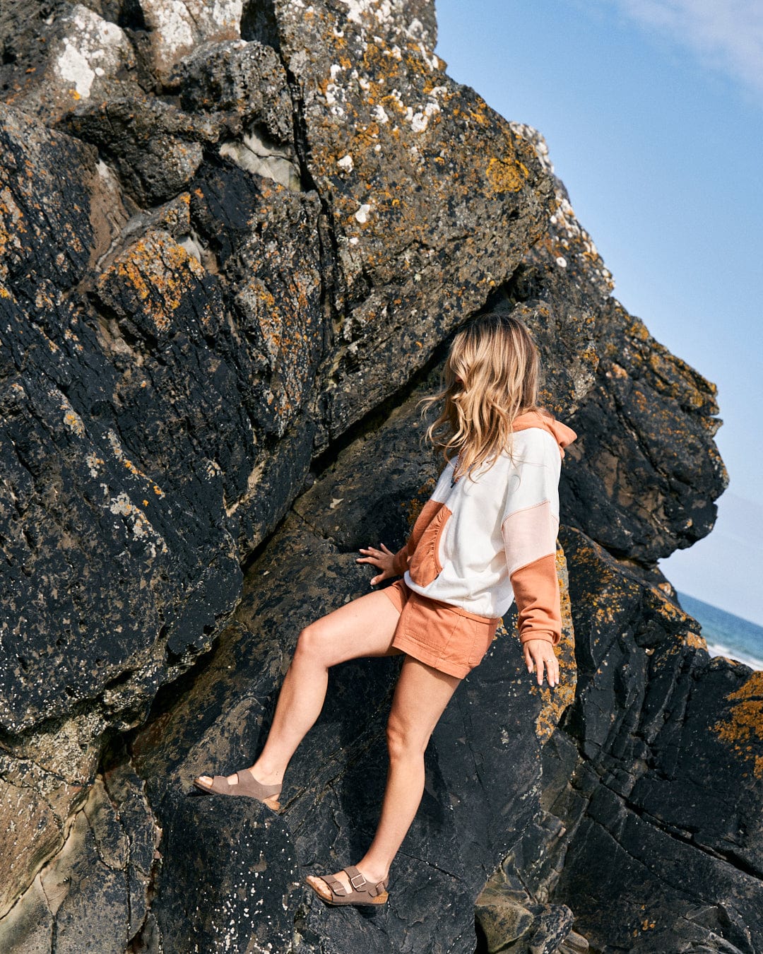 A person in a Palmera - Womens Pop Hoodie - Cream with Saltrock branding and loose-fit shorts climbs on rocky terrain, with the ocean visible in the background.