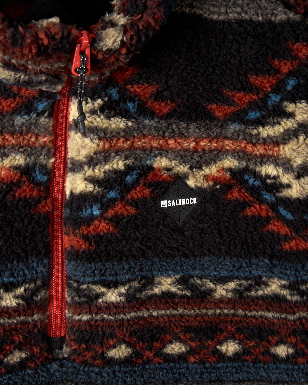 Close-up of the Peninsula Dimensions - Mens Recycled Oversized Fleece by Saltrock, featuring a patterned teddy fleece fabric with a red zipper. The design includes geometric shapes in red, blue, cream, and black colors and is renowned for its cozy oversized fit.