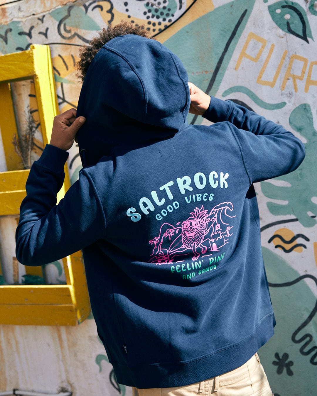 A person wearing a Pine And Sandy - Mens Recycled Zip Hoodie in blue by Saltrock, featuring "Saltrock Good Vibes" with a pink design on the back, stands in front of a colorful, graffiti-style wall.