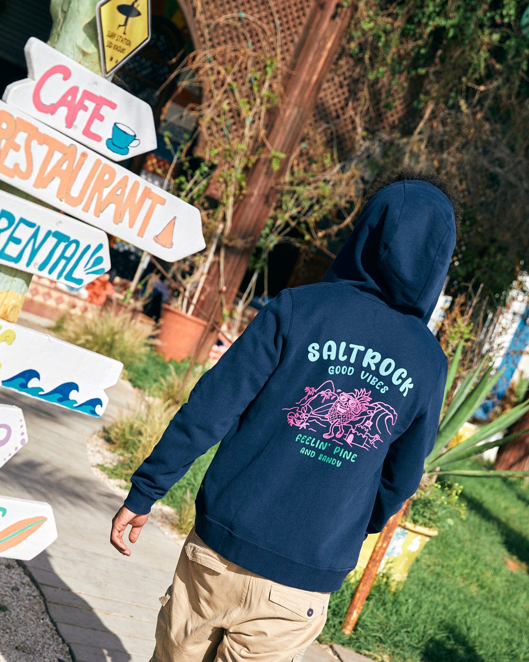 Person wearing the Pine And Sandy - Mens Recycled Zip Hoodie in Blue by Saltrock, which features "Saltrock Good Vibes" and a surfing design on the back, walking past colorful signs pointing to a cafe, restaurant, and rental.