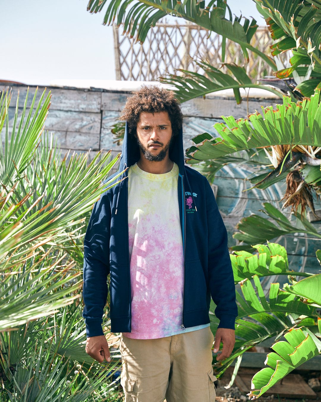 A man stands outdoors surrounded by tropical plants, wearing a tie-dye shirt, khaki shorts, and a Recycled Zip Hoodie in blue from Saltrock's Pine And Sandy collection.