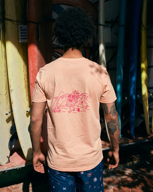A person with curly hair stands with their back to the camera, wearing the Pine And Sandy men's T-shirt in peach by Saltrock, which features graphics in a pink print. Colorful surfboards are lined up against the wall in the background.