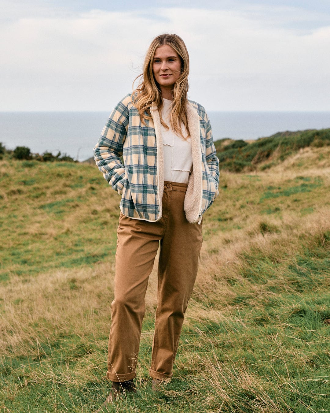 Dressed in the Piper Women's Reversible Zip Collarless Fleece in cream by Saltrock, paired with tan pants and hands in pockets, a person stands on the grassy hillside with the ocean stretching out in the background.