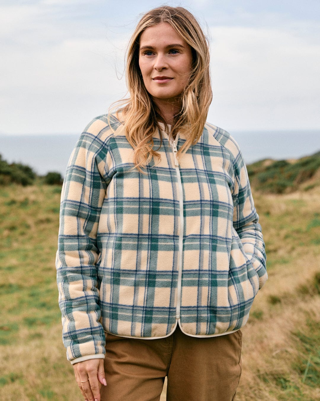 A person stands in a grassy field wearing the Saltrock Piper Women's Reversible Zip Collarless Fleece in cream and khaki pants, with a calm expression.