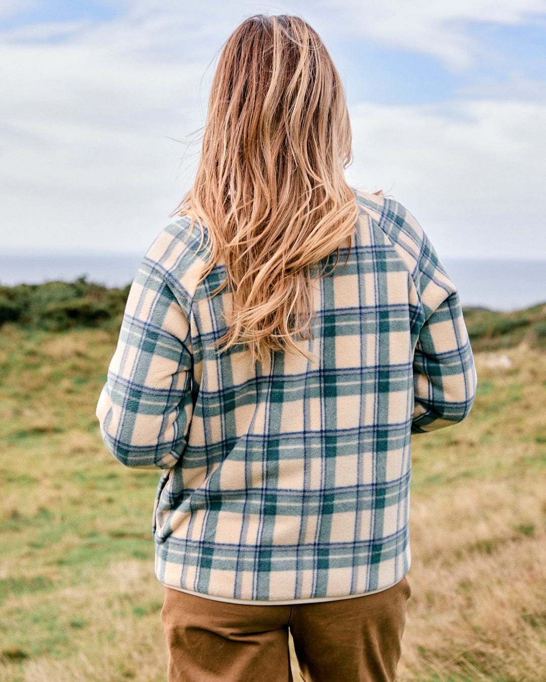 A person with long hair stands outdoors, facing away, wearing the Cream Piper Women's Reversible Zip Collarless Fleece by Saltrock, showcasing eco-friendly materials.