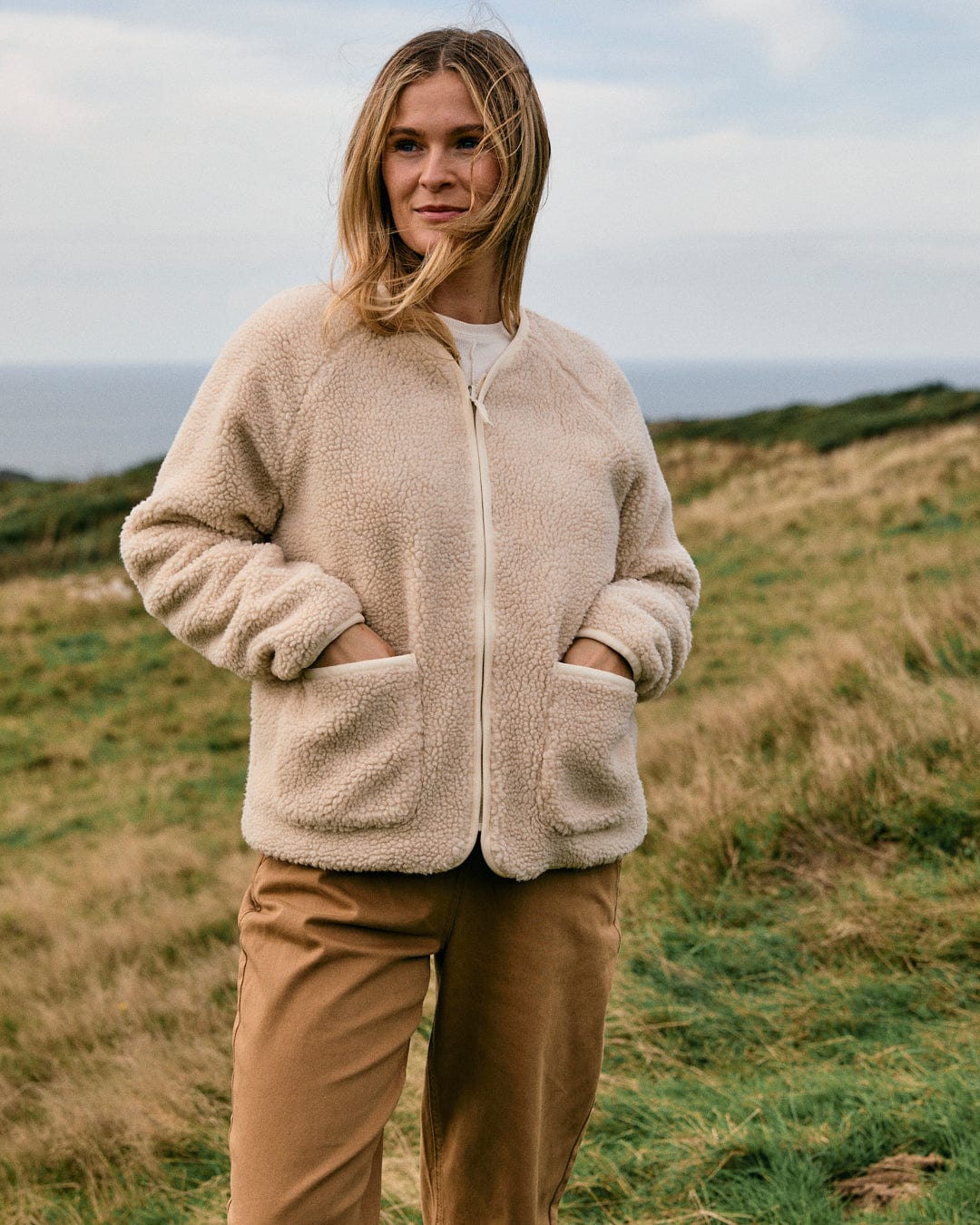 A person wearing the Saltrock Piper Women's Reversible Zip Collarless Fleece in cream stands outdoors on a grassy hillside under an overcast sky.