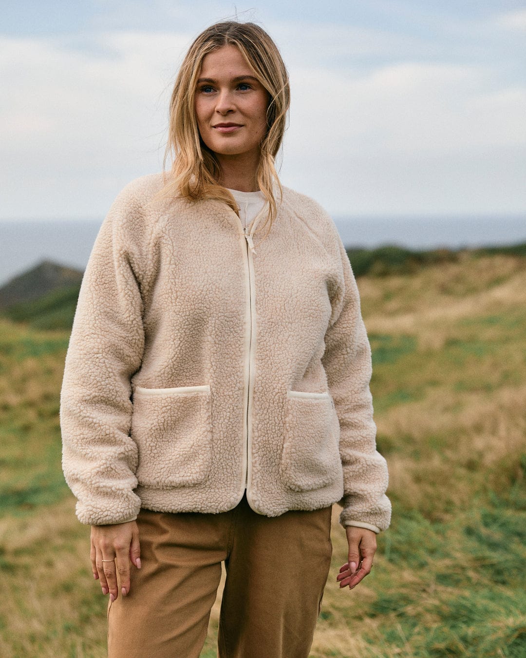 A person stands outdoors on a grassy hill, draped in the Saltrock Piper Women's Reversible Zip Collarless Fleece in cream and light brown pants, framed by the cloudy sky in the background.