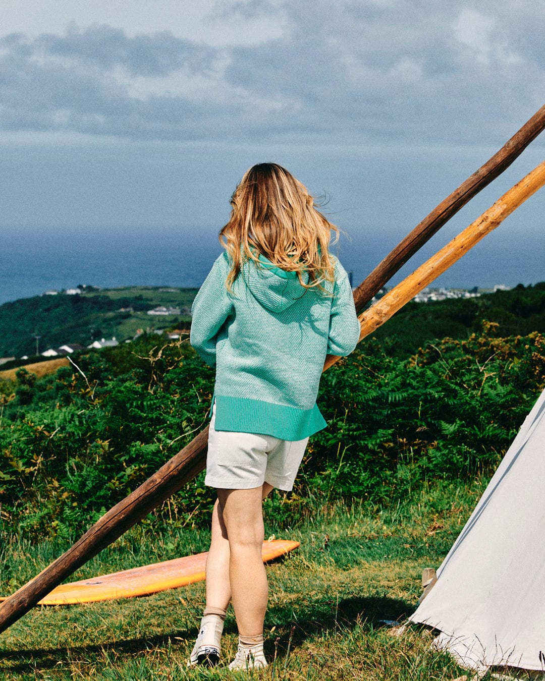 A person with long blond hair stands facing away, wearing the Poppy - Womens Knitted Pop Hoodie in green from Saltrock and white shorts, near a tent in a grassy area with hills and ocean visible in the background.