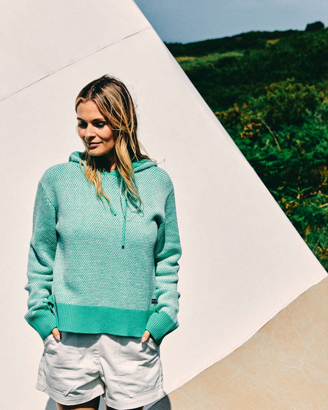 A woman wearing the Saltrock Poppy Women's Knitted Pop Hoodie in green and white shorts stands in front of what appears to be a tent, with greenery in the background.