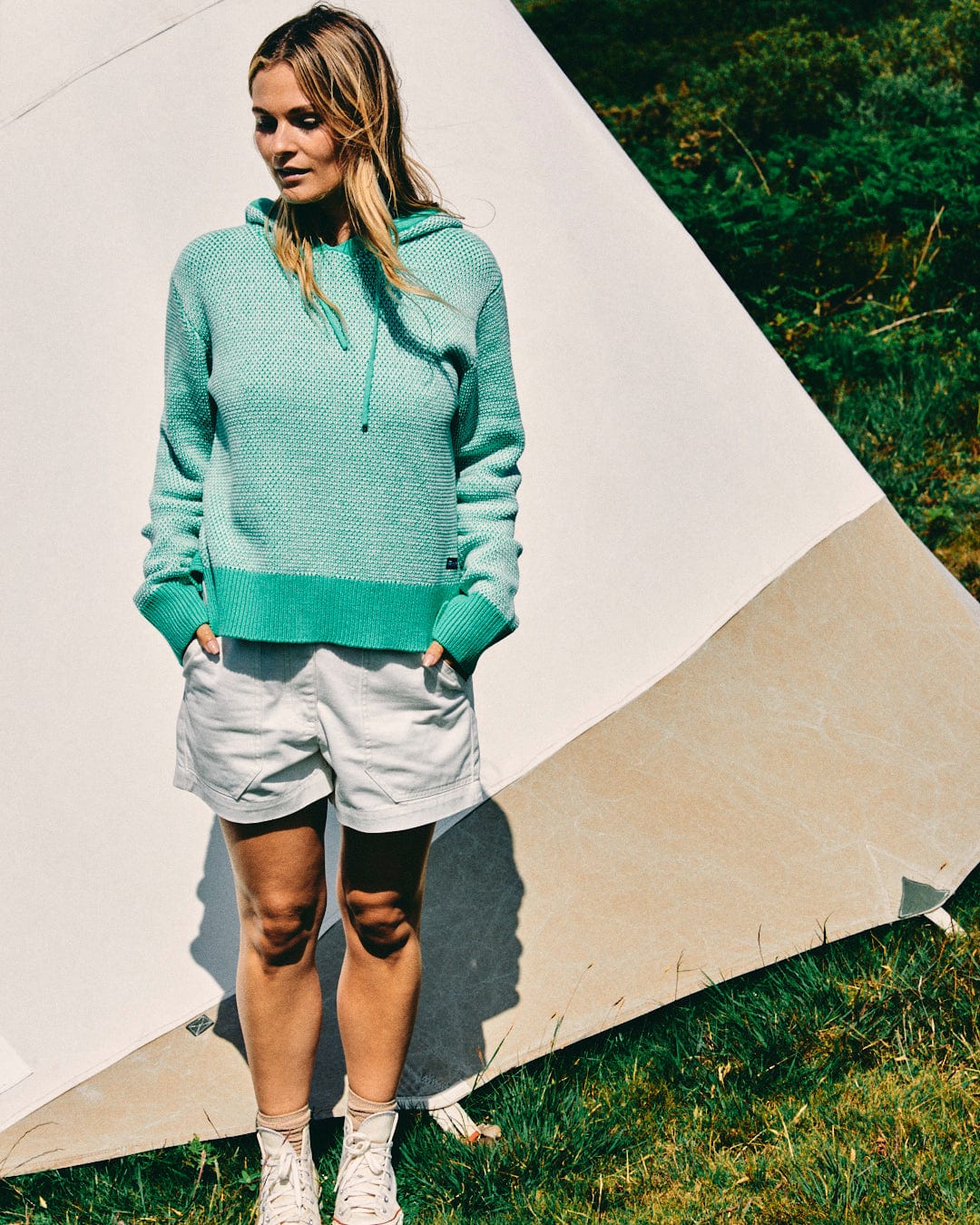 A person stands outside a tent wearing a Saltrock Poppy Women's Knitted Pop Hoodie in green, light-colored shorts, and white sneakers, looking slightly to the side.