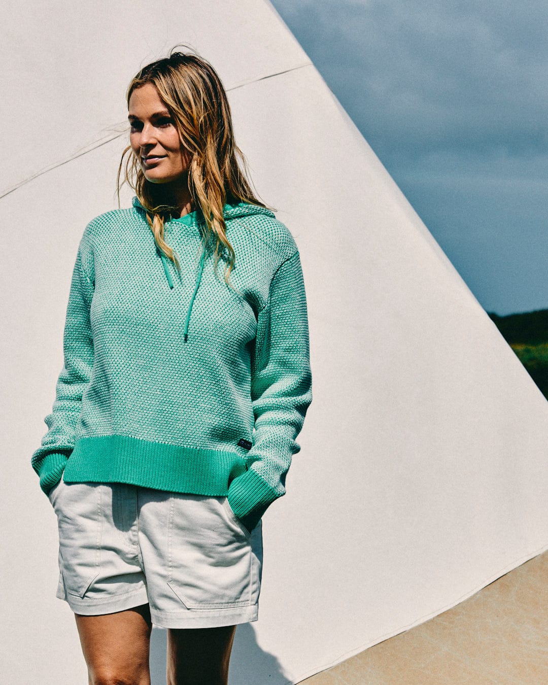 A woman in a Poppy Women's Knitted Pop Hoodie by Saltrock and white shorts stands outdoors with her hands in her pockets.