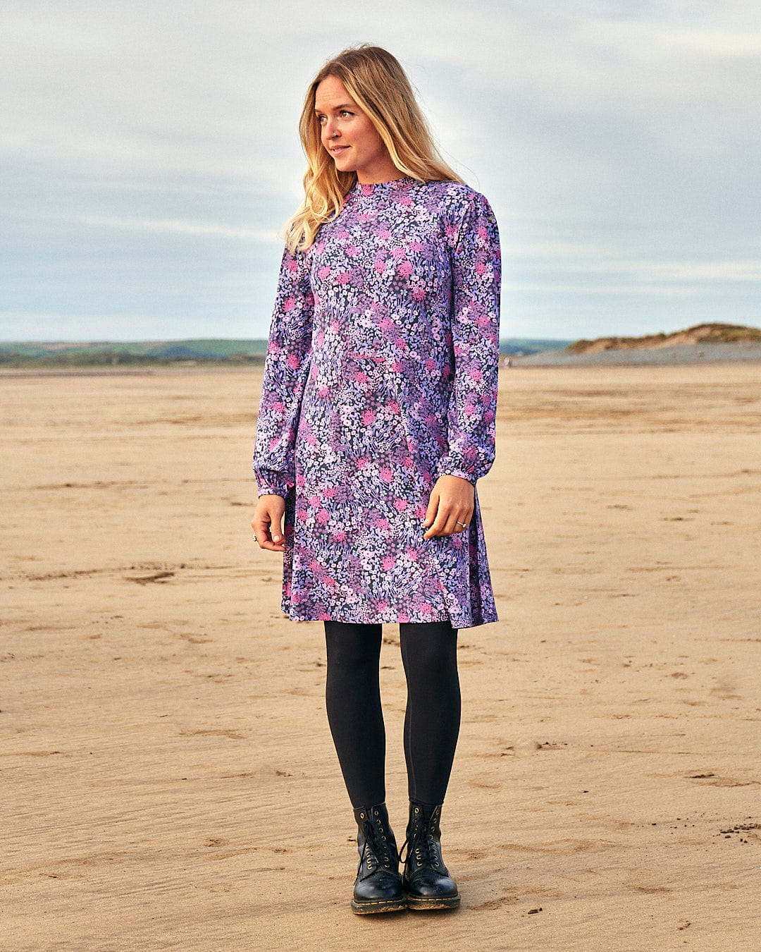 A woman in a Saltrock Ramona dress, a purple floral jersey dress, standing on the beach.