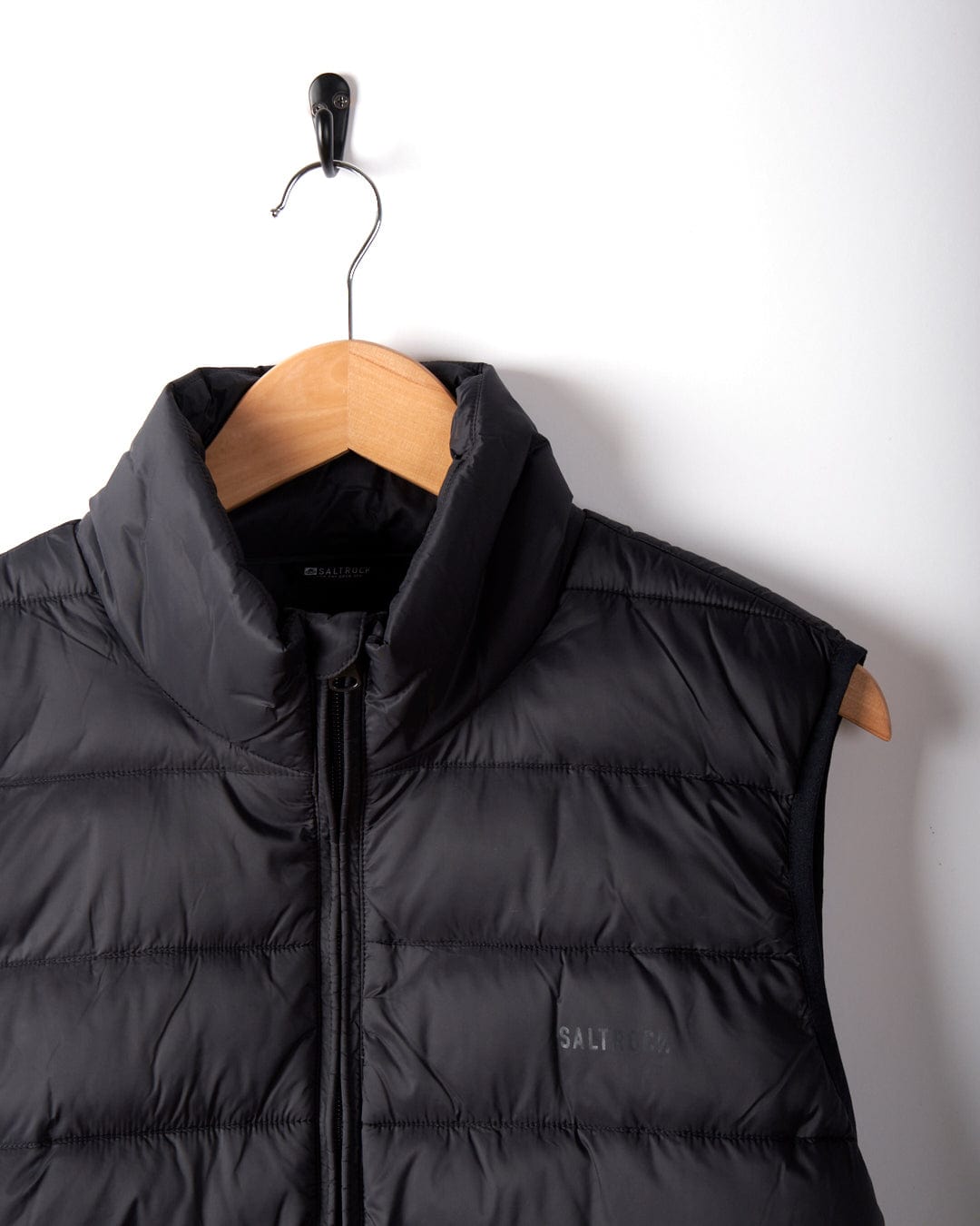 A dark grey Resin Gilet, made from recycled materials and subtly branded by Saltrock, is elegantly draped on a wooden hanger from a wall hook.