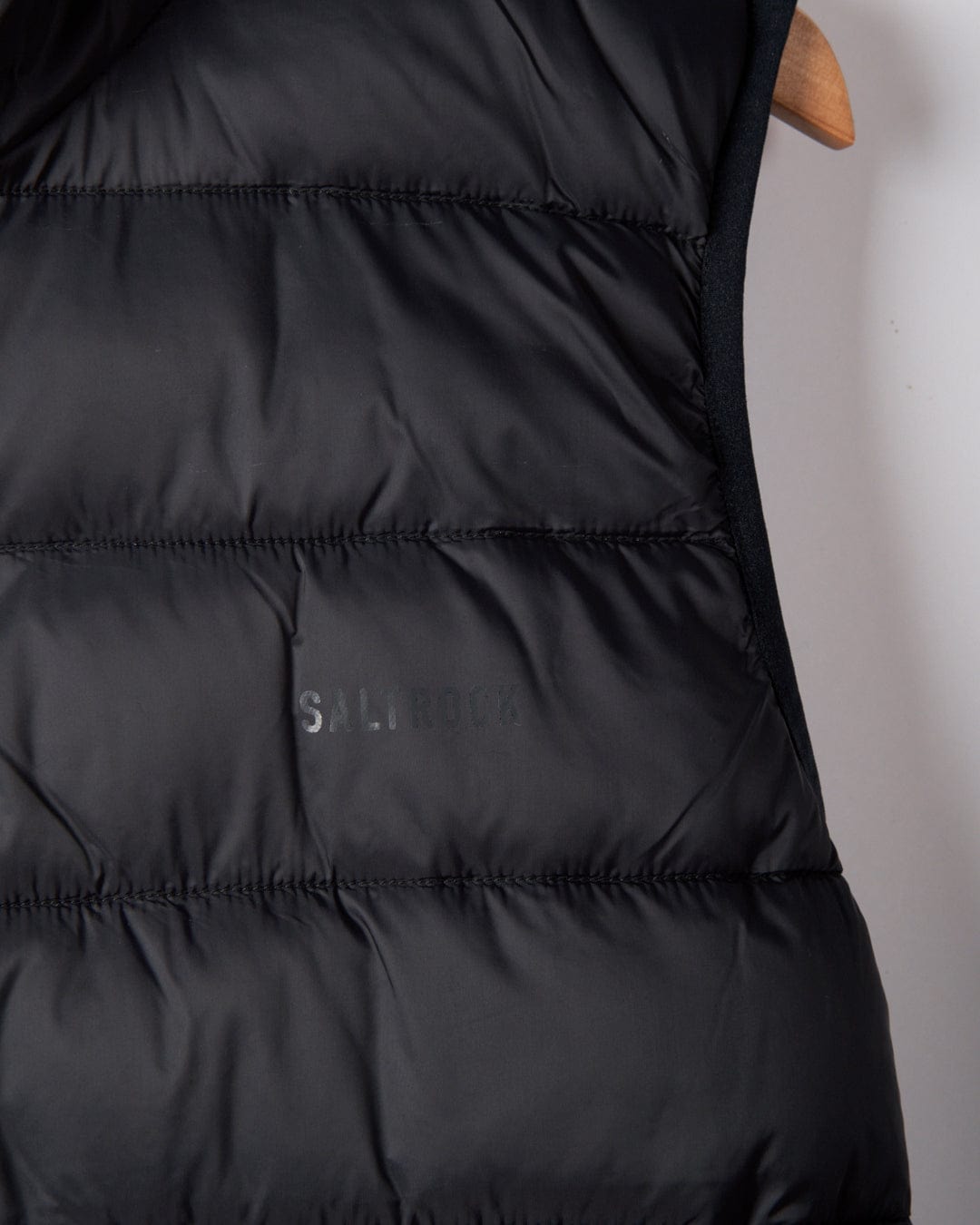 Close-up of a dark grey quilted Saltrock Resin Gilet, crafted from recycled materials and water-resistant fabric, hanging on a wooden hanger; the "S--ROCK" branding partially visible.