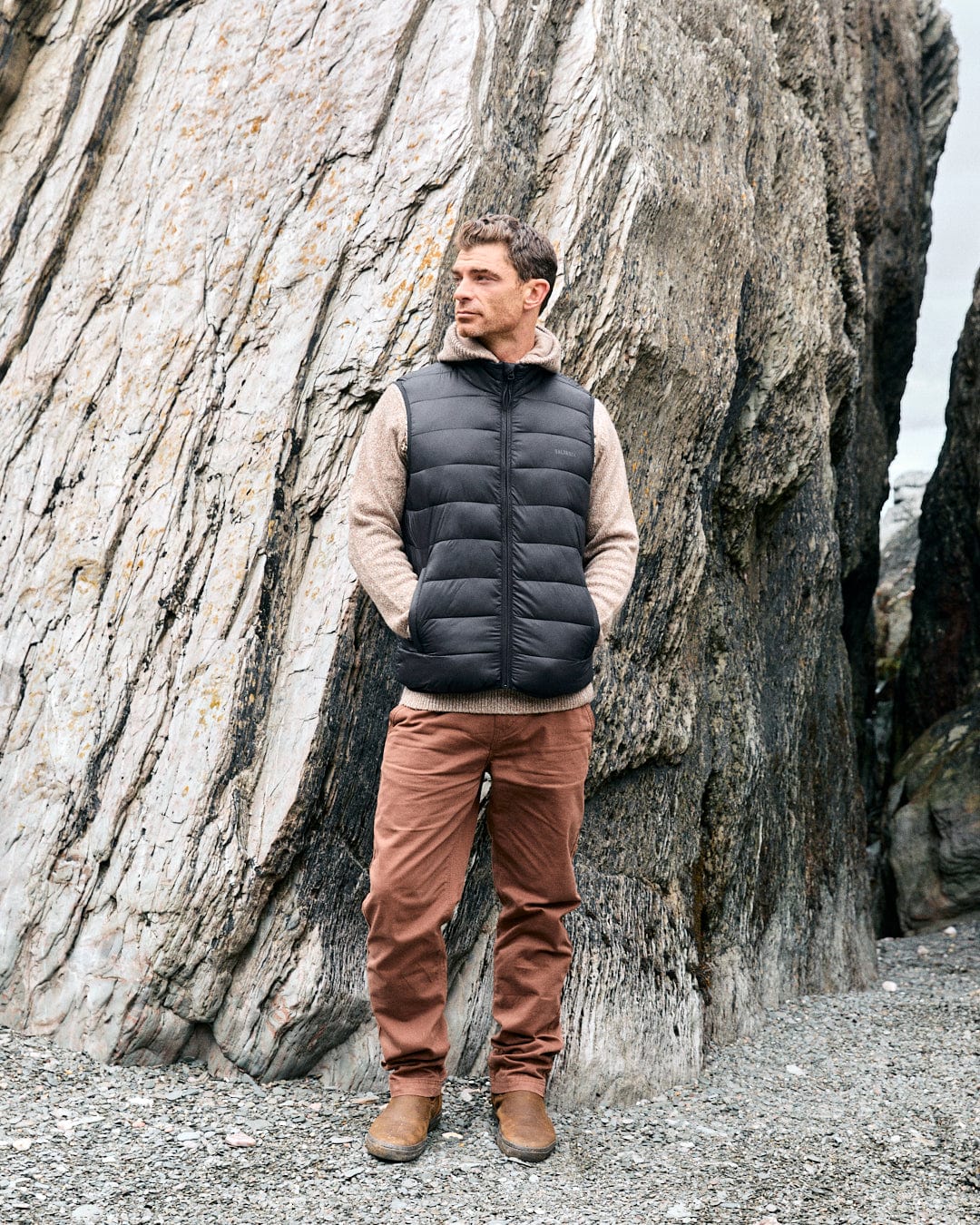 A man in a water-resistant, dark grey Resin Gilet by Saltrock, a beige sweater, and brown pants stands on rocky terrain beside large rock formations.