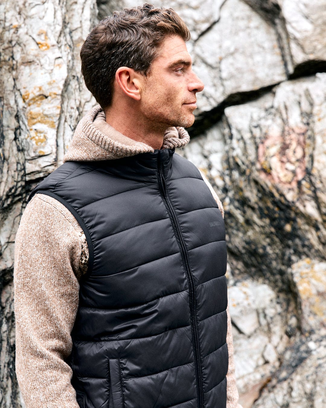 A man in a dark grey Resin Gilet by Saltrock, made from recycled materials, stands sideways against a rocky backdrop.