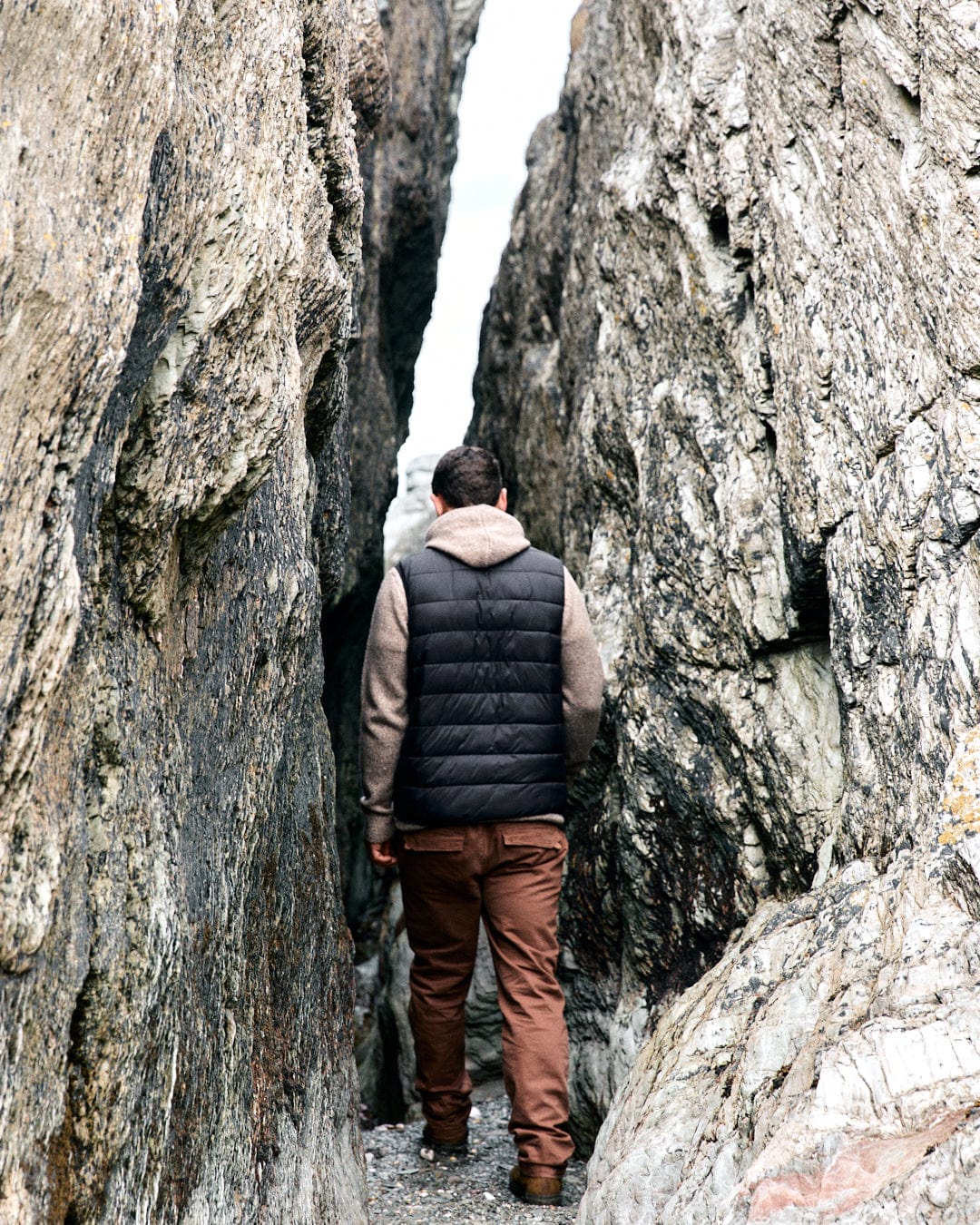 Wearing a water-resistant dark grey Resin Gilet and brown pants, a person strides confidently between tall rock formations, embodying the rugged style of Saltrock.