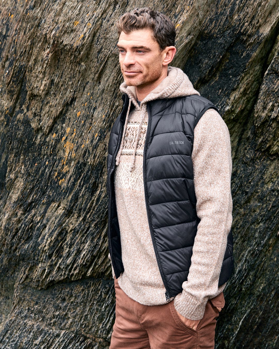 A man wearing Saltrock's Resin Gilet, a dark grey men's gilet made from recycled materials, stands against a textured rock background, gazing to the side.