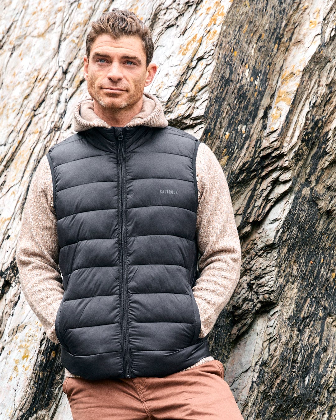 A man wearing the Saltrock Resin Gilet - Mens Gilet in Dark Grey and a beige hoodie stands against a rocky backdrop, looking to the side.