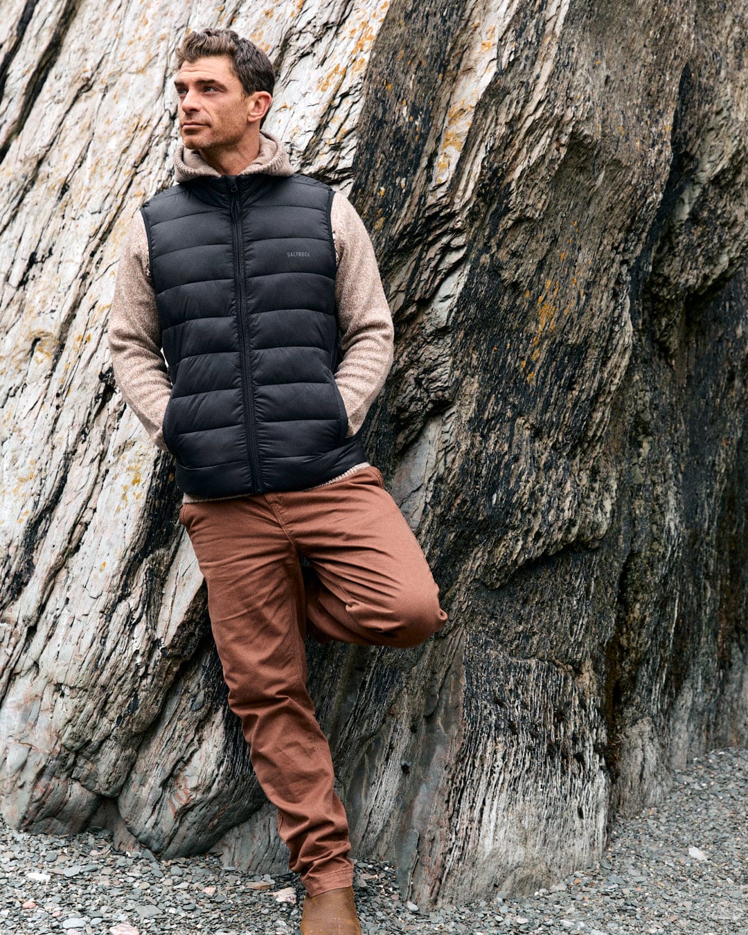 A man leans on a rocky surface, wearing a Saltrock dark grey Resin Gilet crafted from recycled materials. Complementing his look are a beige hoodie, brown pants, and sturdy Saltrock-branded boots.