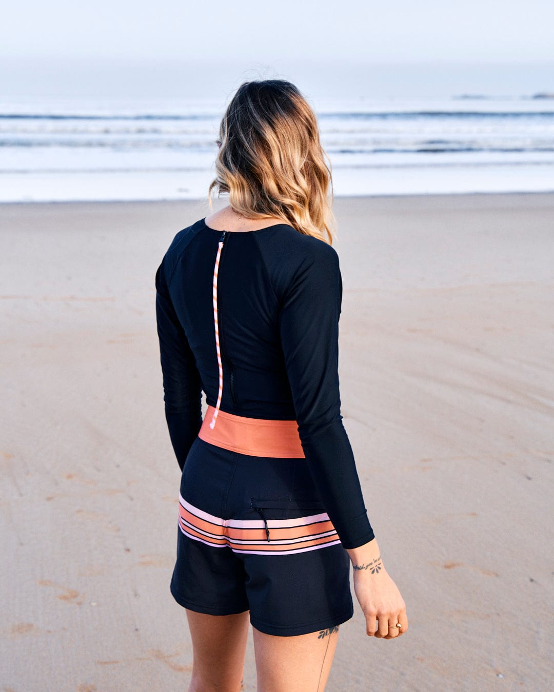 A person with long hair stands on a beach facing the ocean, wearing a black long-sleeve top and Saltrock Retro Ribbon - Womens Boardshorts - Black with pink accents made from recycled material.