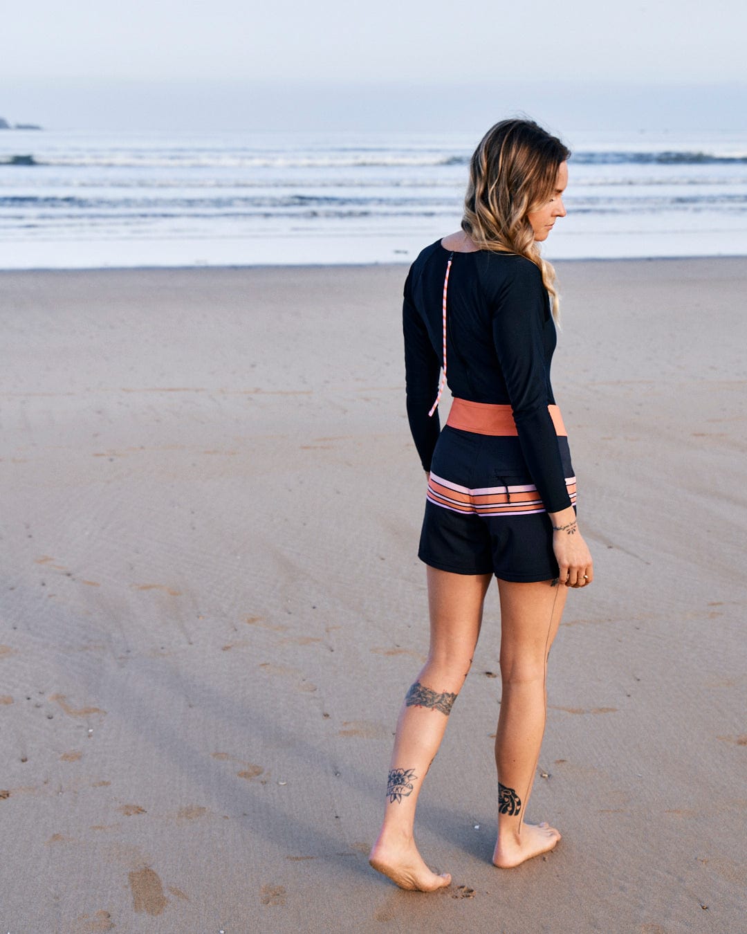 A person with long hair walks barefoot on the beach, looking towards the ocean. They wear Saltrock's Retro Ribbon - Womens Boardshorts - Black, made from recycled materials, and have tattoos on their legs. The sky is clear and the water is calm.