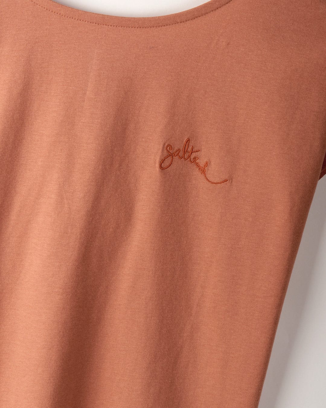 Close-up of a rust-colored, 100% cotton t-shirt with the word "Saltrock" embroidered on the chest in a cursive font and dropped back detail.