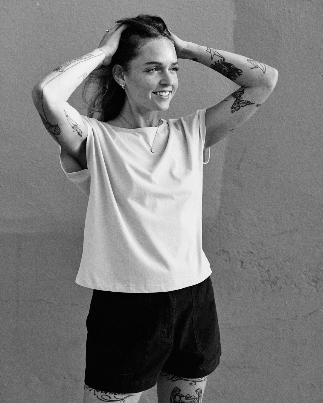 A person with tattoos, wearing a Saltrock Rhoda - Womens T-Shirt - Light Blue and dark shorts, stands against a wall. They are smiling and have their hands on their head. The shirt, made from 100% cotton, features a dropped back detail that adds a unique touch to the casual outfit.