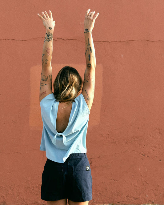 Person with tattooed arms, seen from the back, wearing a sleeveless Saltrock Rhoda - Womens T-Shirt - Light Blue with a dropped back detail and dark shorts, raising their arms against a textured pink wall.