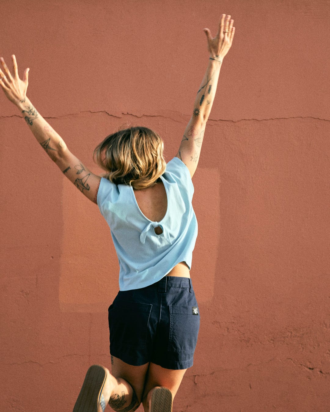 A person wearing a light blue, 100% cotton Rhoda - Womens T-Shirt - Light Blue by Saltrock with a dropped back detail and dark shorts is jumping with their arms raised against a pinkish wall. Tattoos are visible on both arms.