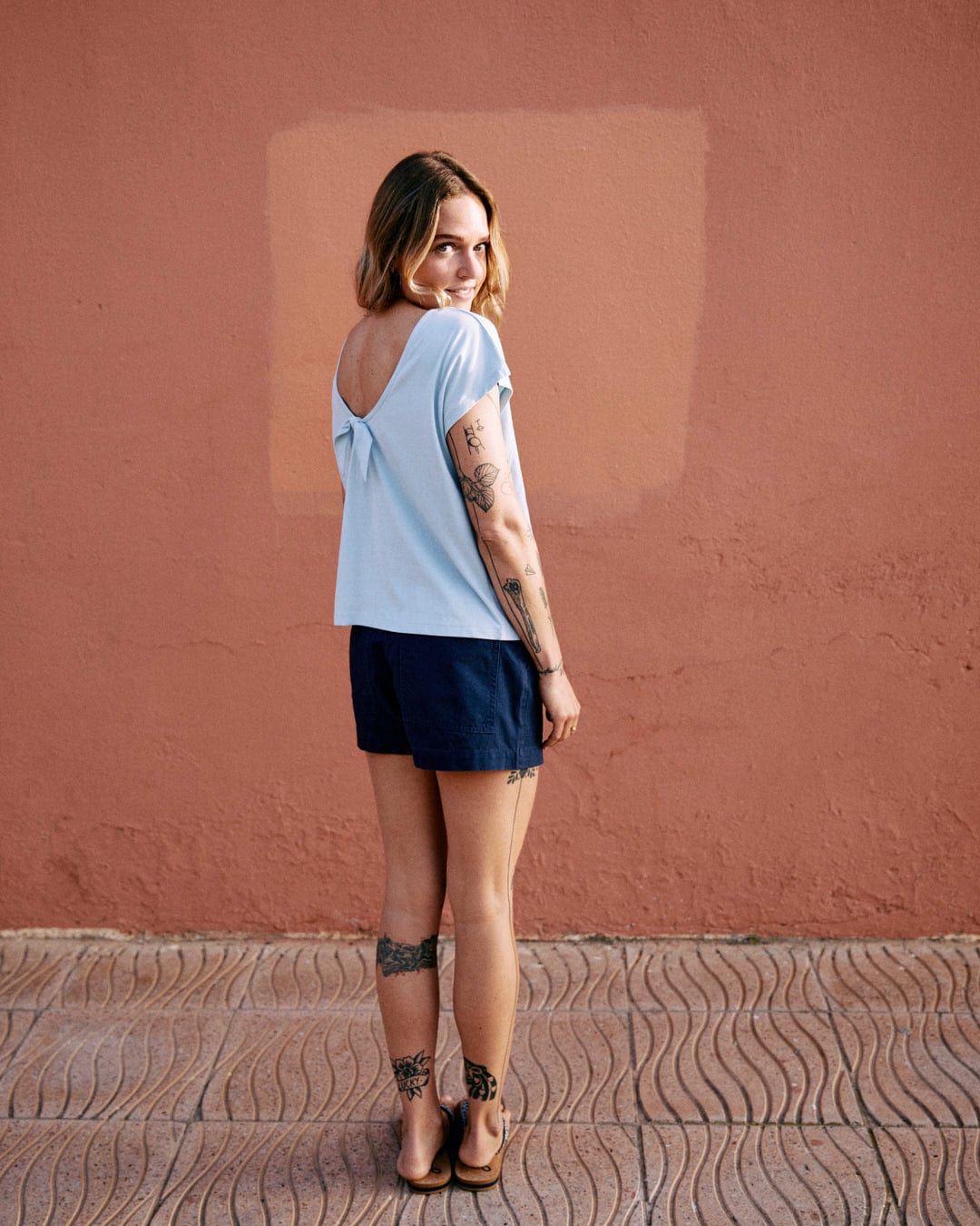 A woman with tattoos wearing a Saltrock Rhoda - Womens T-Shirt - Light Blue featuring a dropped back detail and navy shorts is standing outdoors against a pink wall, looking back over her shoulder and smiling.