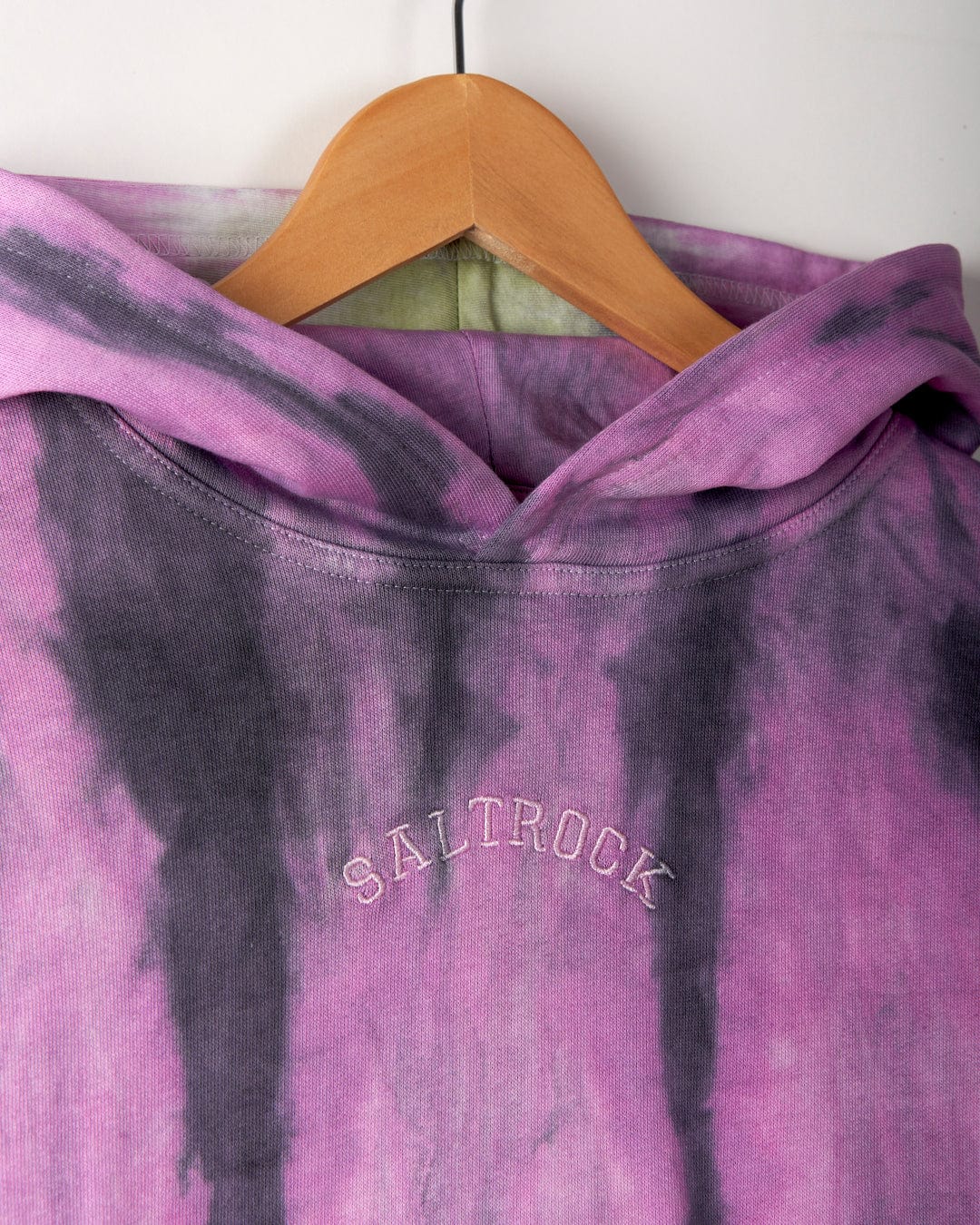 Close-up of a purple and black tie-dye hoodie on a wooden hanger, featuring the Saltrock branding with embroidered text "SALTROCK" on the back. Made from 100% cotton, this Rock Valley - Pop Hoodie - Multi showcases a vibrant tie dye print that stands out.