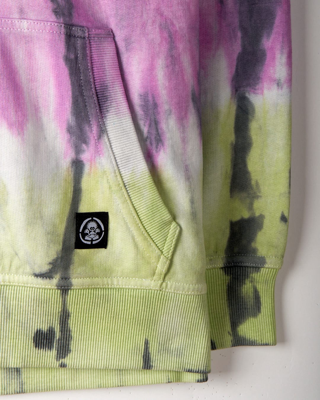 Close-up of a tie-dye print hoodie with pink, purple, green, and black patterns, crafted from 100% cotton. It features a small Saltrock branding logo with a skull and crossbones near the pocket. The product is the Rock Valley - Pop Hoodie - Multi by Saltrock.