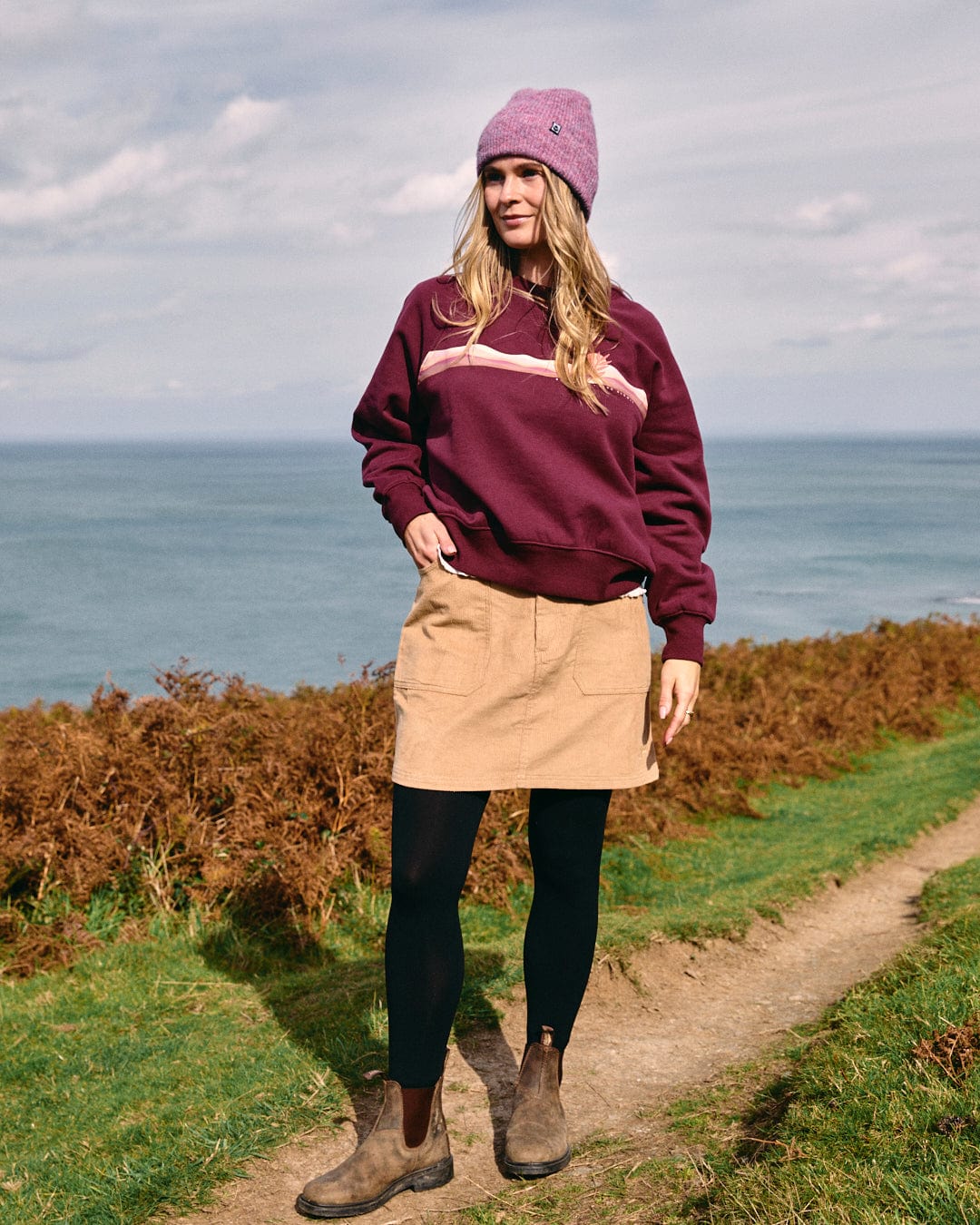 A person stands on a grassy path by the sea, wearing a maroon sweater, black leggings, brown boots, and a purple beanie, complemented by the Sadie women's cord skirt in brown from Saltrock.