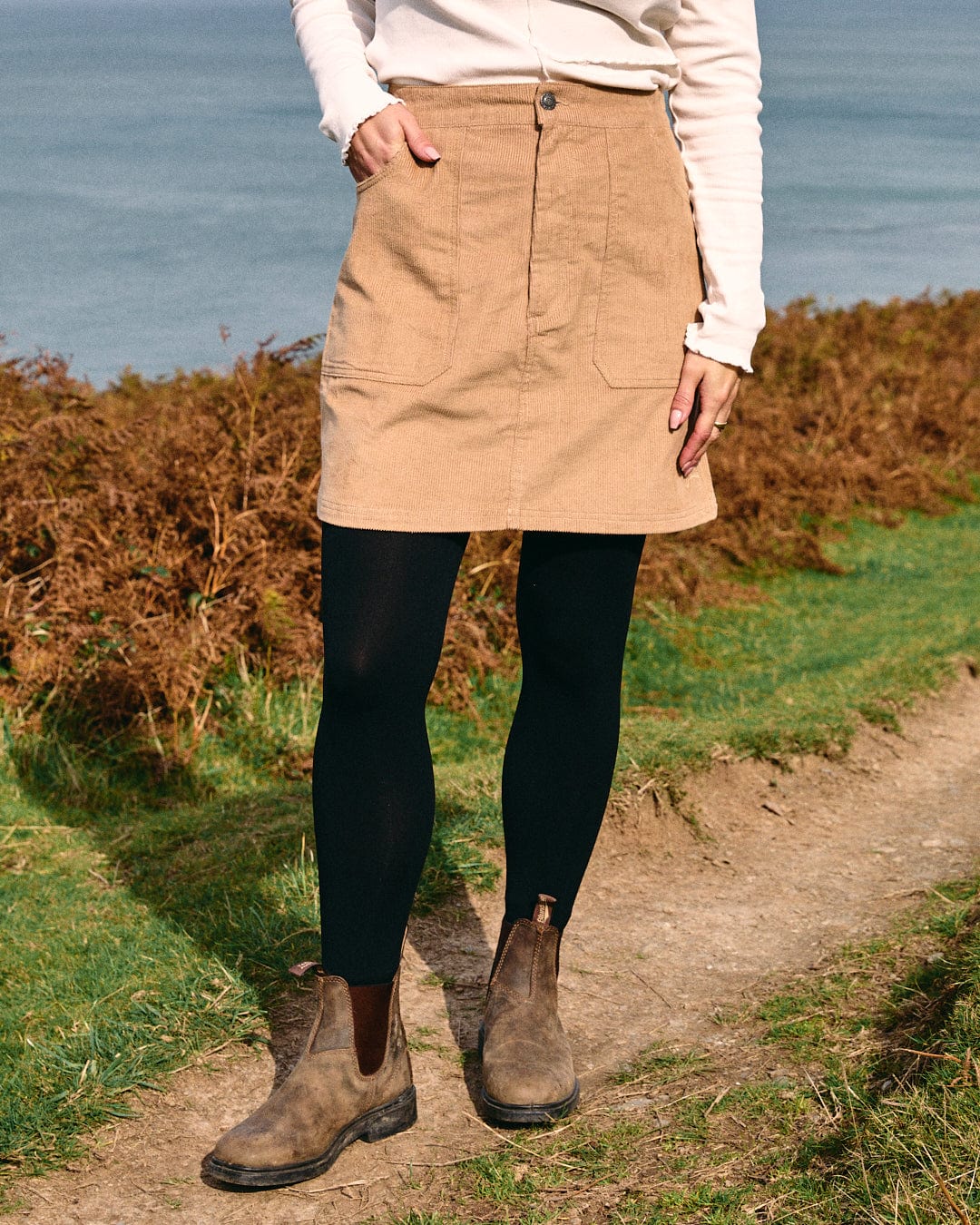 A person stands on a grassy path by the sea, wearing Saltrock's Sadie women's cord skirt in brown of mini length, paired with black tights, brown boots, and a crisp white top.