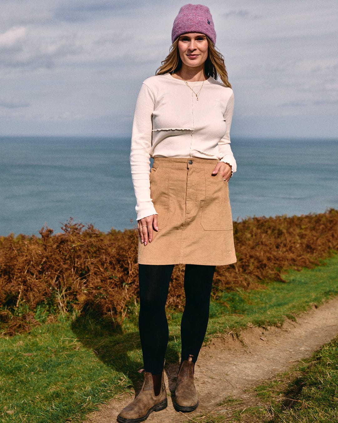 A person stands on a grassy path with the ocean in the background, wearing a white top, the Saltrock Sadie - Women's Cord Skirt in brown of mini length, black leggings, brown boots, and a purple beanie.