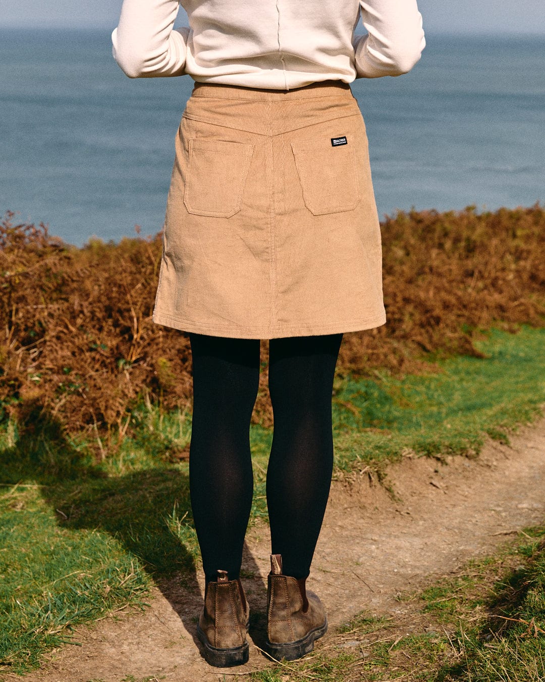 A person in a brown Sadie cord skirt from Saltrock, paired with black tights and brown boots, stands on a grassy path overlooking the ocean.