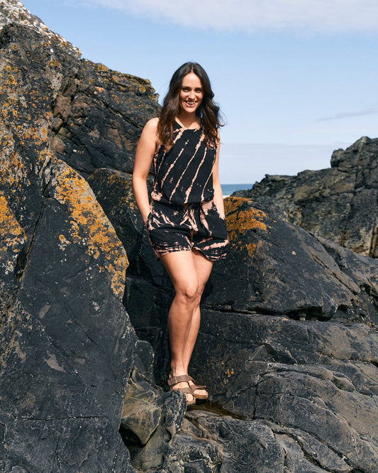 A woman in a lightweight material Saltrock Saffron - Womens Tie Front Vest - Black stands on jagged rocks outdoors, smiling at the camera.