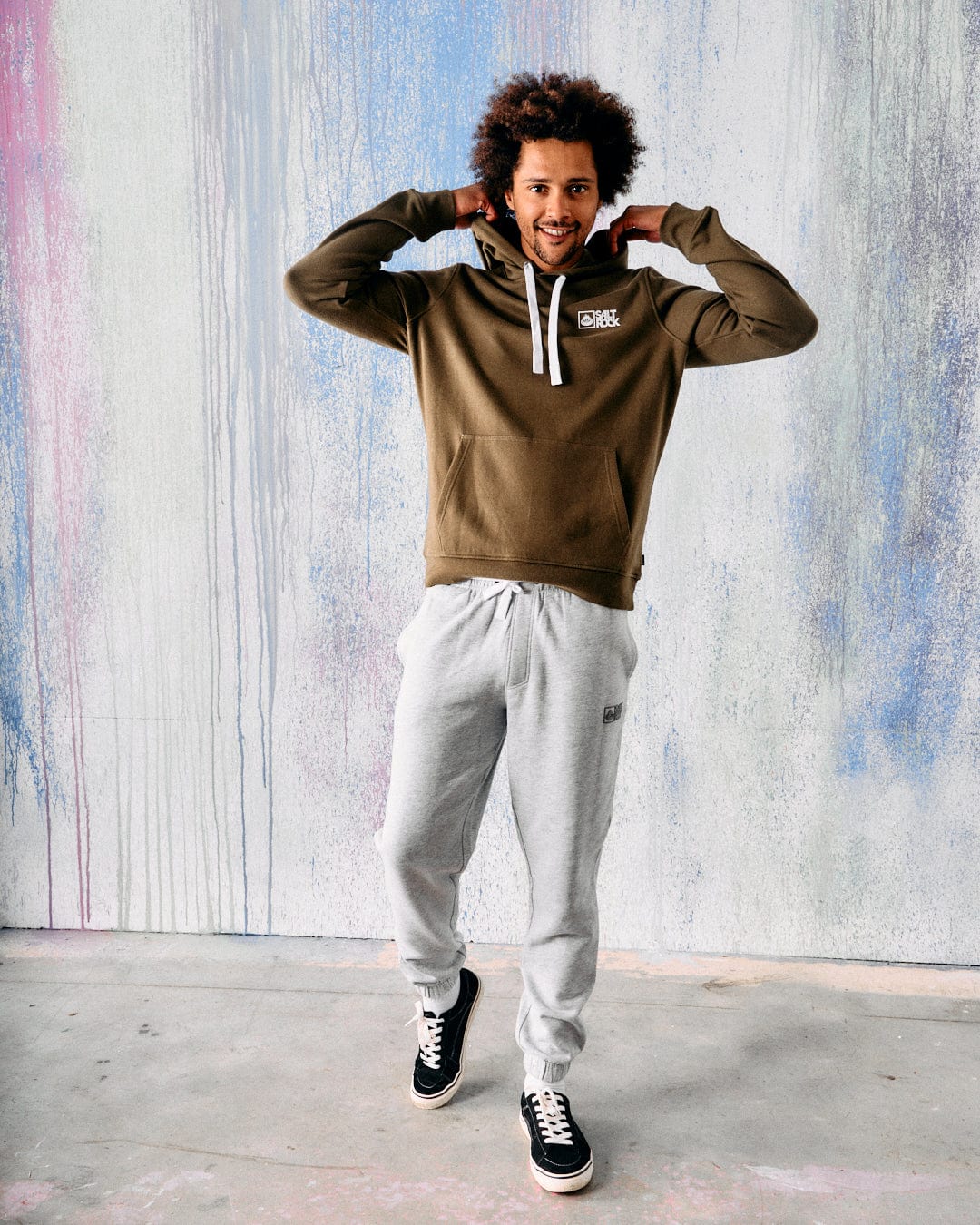 A person smiles in a cozy green Saltrock Original hoodie with a drawcord hood, paired with gray sweatpants. They stand on a concrete floor against a textured multicolored wall. The soft jersey material and subtle Saltrock branding enhance comfort and style.