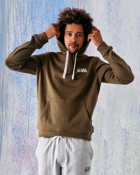 A man adjusts his Saltrock Original - Mens Pop Hoodie in green, featuring the Saltrock branding and a white logo, crafted from soft jersey with a drawcord hood for comfort. He poses against a blue and white textured backdrop, paired with light gray sweatpants.