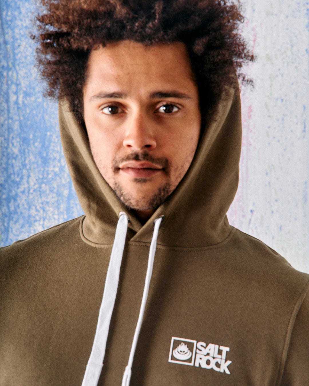 In front of a textured blue and white background, someone with curly hair wears the Saltrock Original Men's Pop Hoodie in green, showcasing the Saltrock brand and made from soft jersey material.