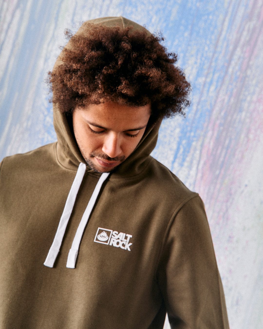 A person with curly hair is wearing a green Saltrock Original men's pop hoodie crafted from soft jersey material, complete with white drawstrings and the signature Saltrock branding. They are looking downwards against a pastel-colored patterned background.