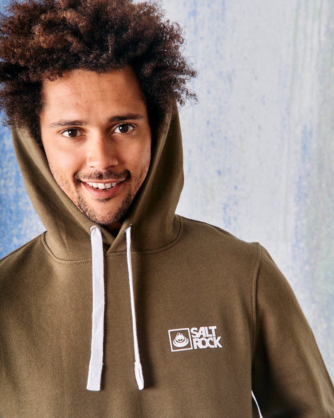 A person with curly hair smiles warmly in their Saltrock Original Men's Pop Hoodie in green, featuring the "Saltrock" logo. Its soft jersey fabric offers comfort, and the drawcord hood adds style against a speckled blue and white backdrop.