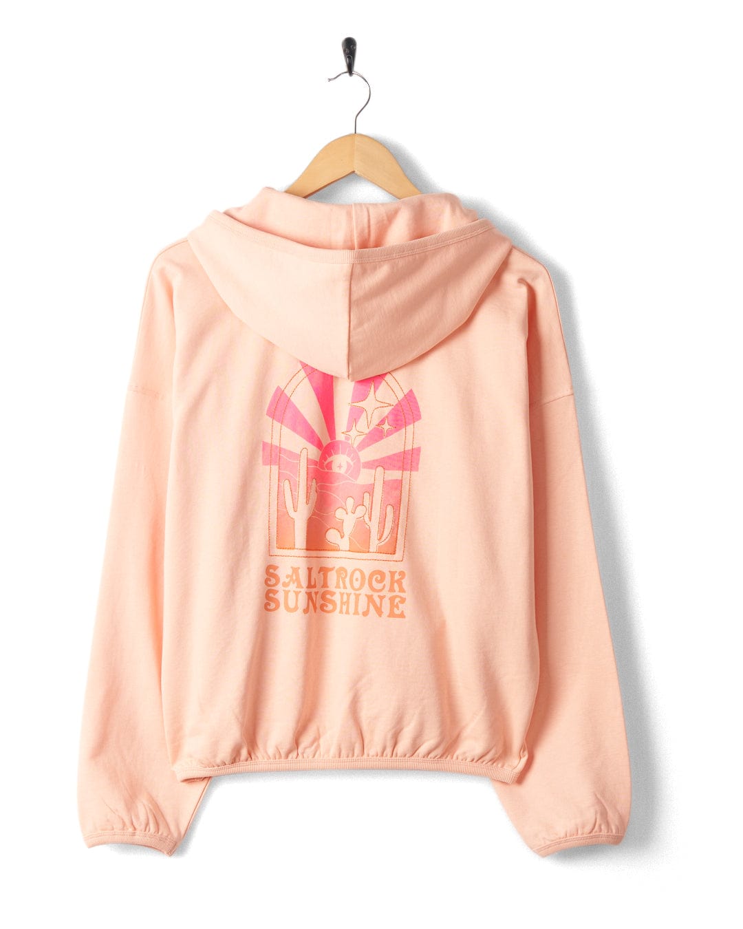 A peach-colored hooded sweatshirt hangs on a wooden hanger, featuring a mystical ombre print of a sunset, surfboards, and cacti with the text "Saltrock Sunshine" embroidered on the back. Made from 100% cotton for ultimate comfort, it is the Saltrock Sunshine - Womens Pop Hood - Peach by Saltrock.