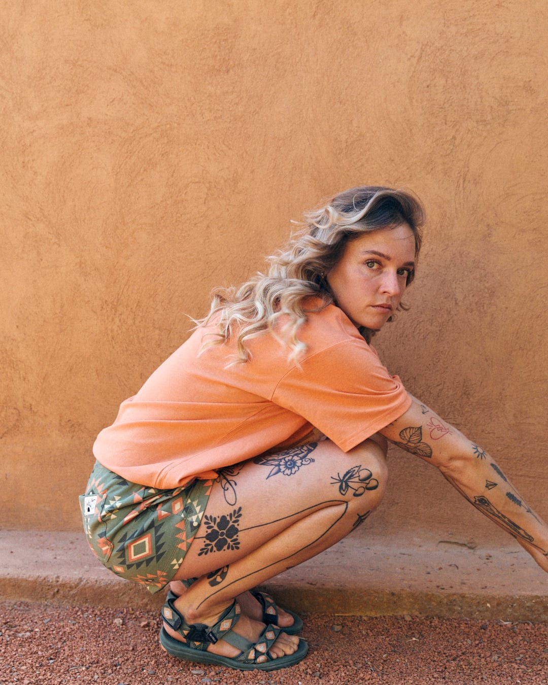 A person with tattoos on their arms and legs, sporting a Saltrock Sunshine - Recycled Womens Cropped T-Shirt - Orange, squats against an orange textured wall. They have shoulder-length wavy hair, patterned shorts, and sandals that add to the Saltrock branding vibe.